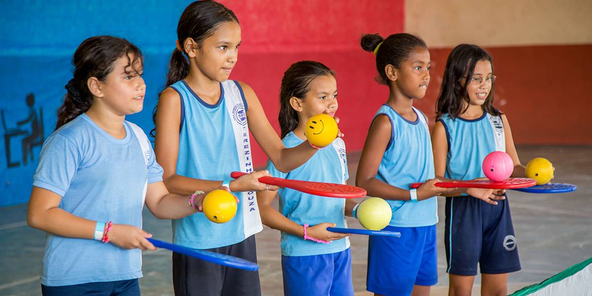Cinco meninas enfileiradas lado a lado seguram raquetes com uma das mãos. Com a outra, elas seguram pequenas bolas.