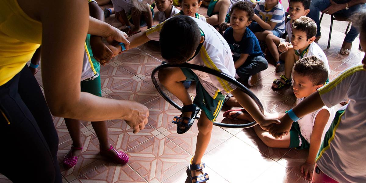 Garoto, de mãos dadas com dois colegas, curva-se para passar sua perna por dentro de um bambolê sem soltar-se dos amigos. Educadora acompanha a atividade ao lado.