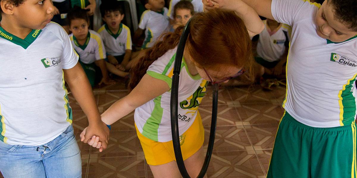 Menina, de mãos dadas com dois colegas, passa bambolê pelo pescoço sem soltar-se dos amigos.