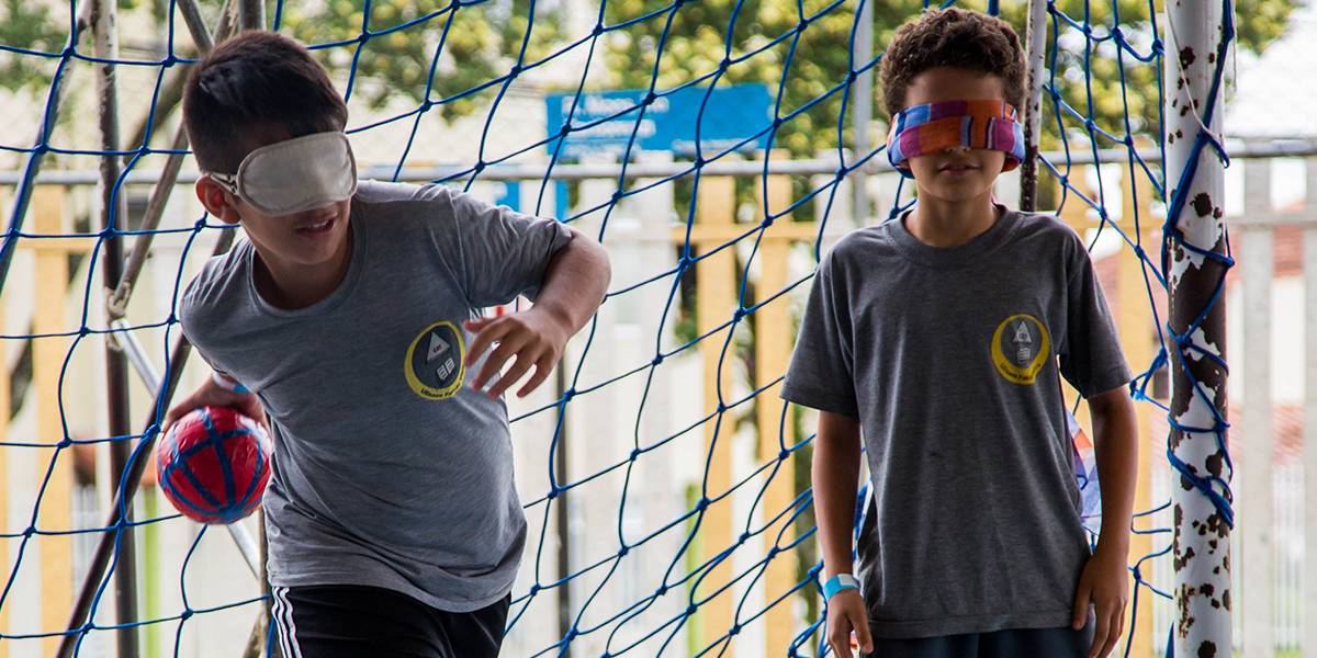 Dois garotos com os olhos vendados jogam golbol. Eles parados de costas para o gol e um deles lança uma pequena bola.