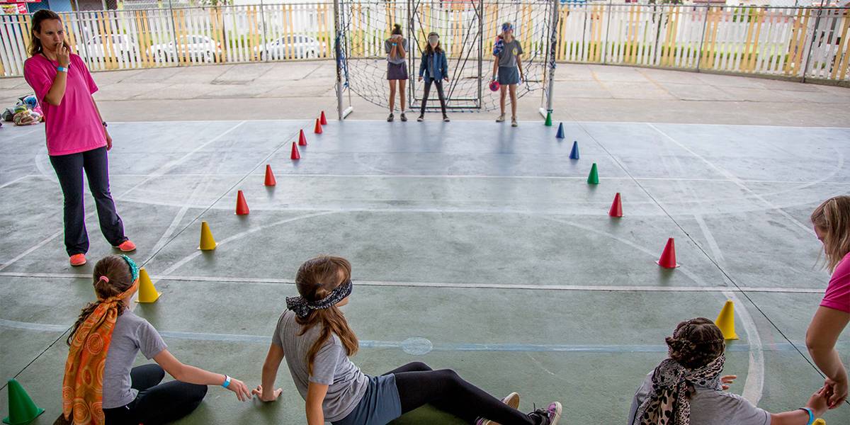 Seis estudantes vendados jogam golbol, em duas equipes de três. Cada time está de um lado da quadra, que é delimitada por pequenos cones coloridos. A professora acompanha a atividade ao lado, fora da marcação.