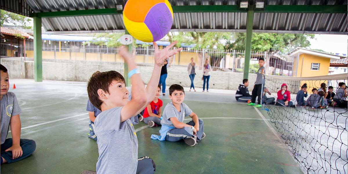 Estudante sentado na quadra joga bola de vôlei sobre a rede para o campo adversário. Atrás, seus colegas de time, também sentados no chão, acompanham a jogada.