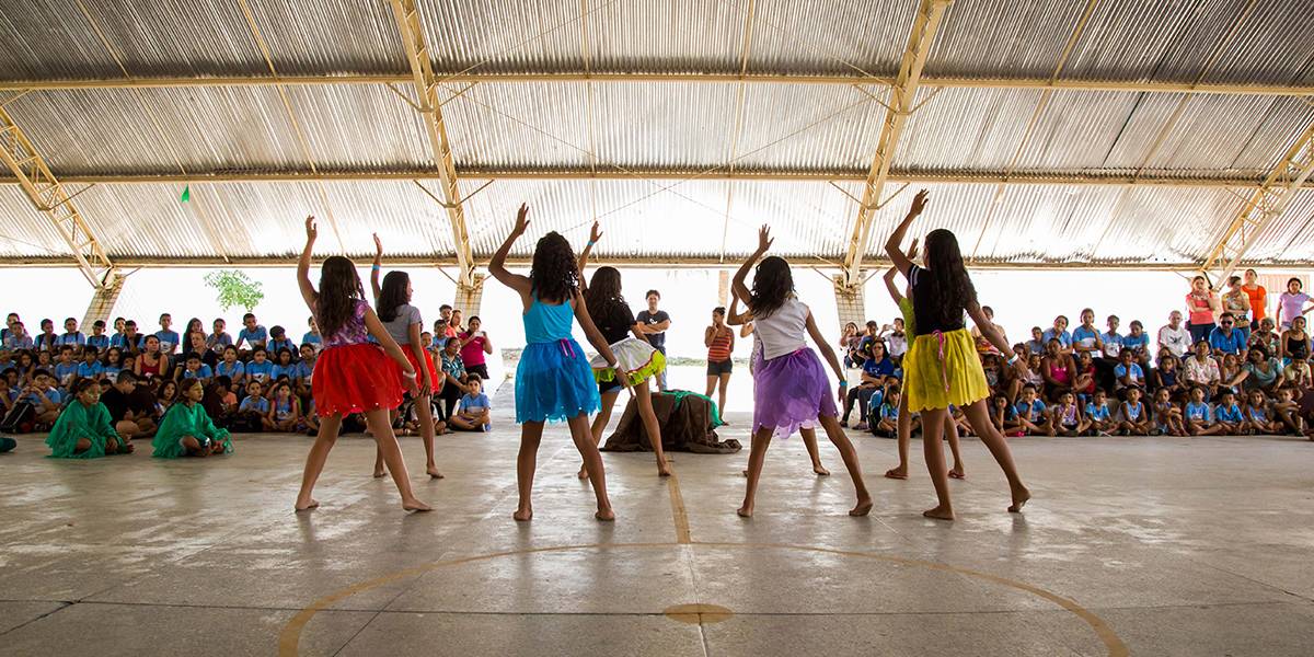 Grupo de garotas com saias coloridas faz coreografia diante de espectadores na arquibancada.