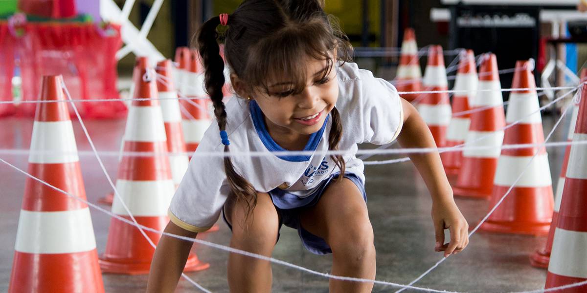 Menina se agacha para passar sob túnel formado por uma trama de barbantes amarrados ao topo de cones.