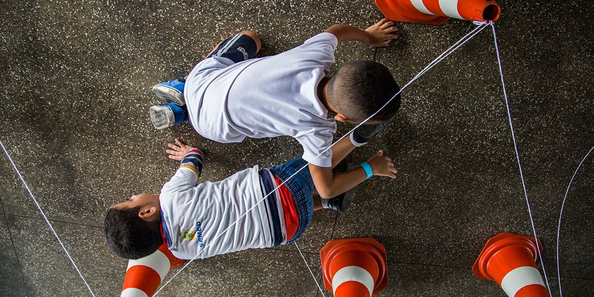 Dois meninos engatinham em sentidos opostos para passar por baixo de barbantes amarrados ao topo de cones.