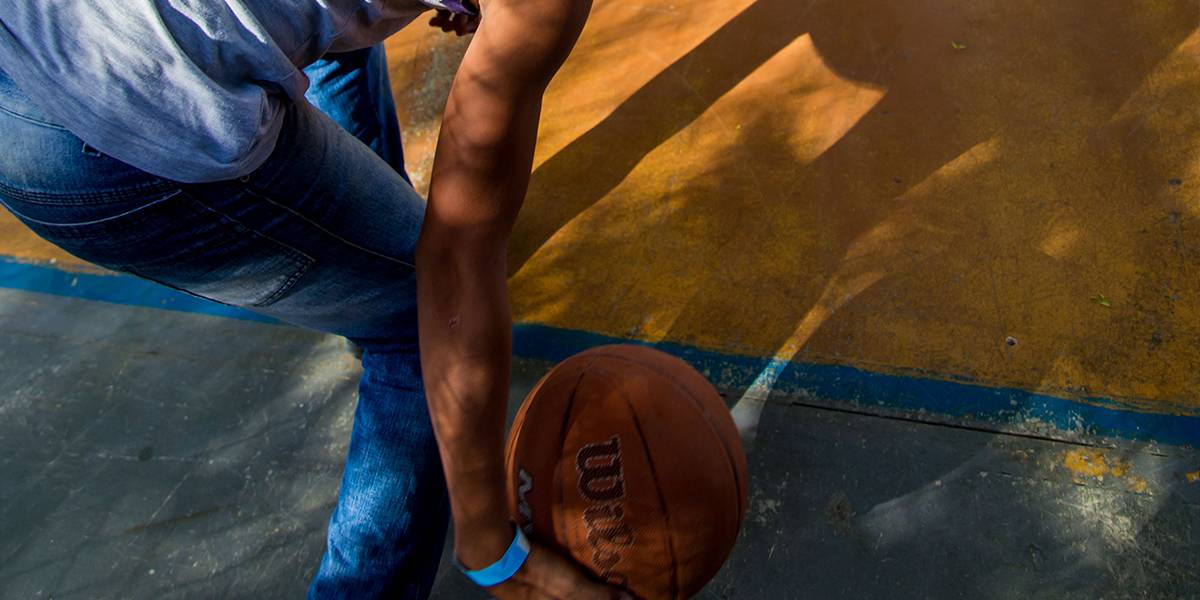 Garoto abaixa-se para lançar bola de basquete no chão em direção a um conjunto de bolas no lado oposto da quadra.