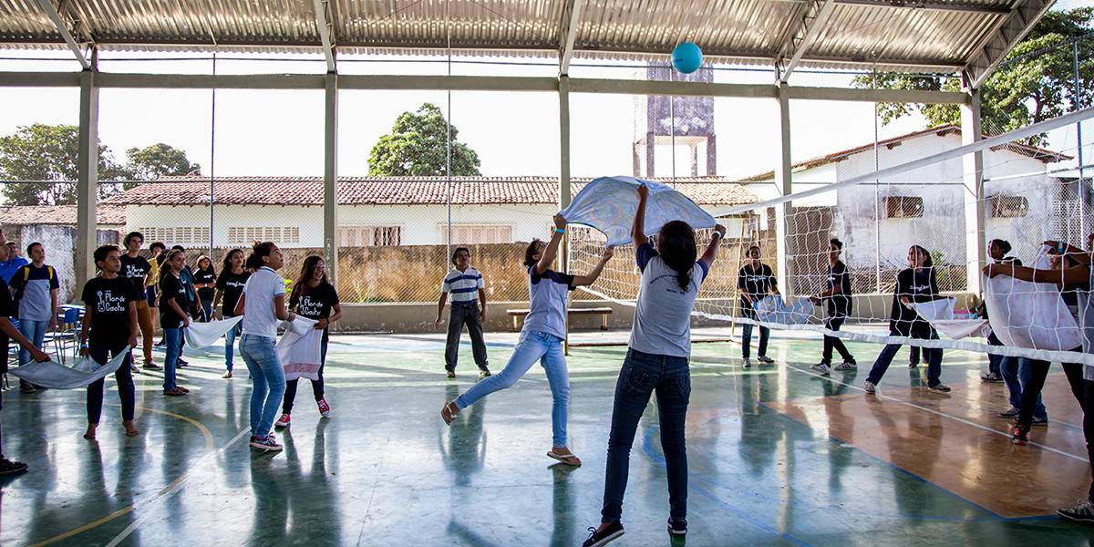 Duas garotas seguram toalha de banho pelas pontas e o usam para lançar bola sobre rede em direção ao lado adversário. Os jogadores dos dois times acompanham a jogada, segurando suas toalhas em duplas.