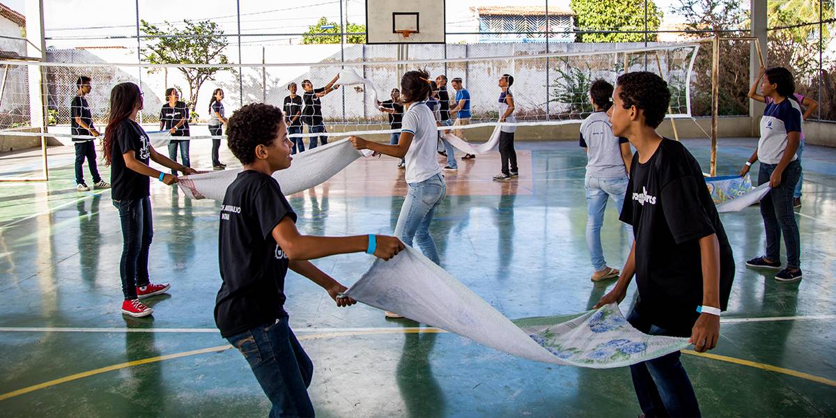 Dois times de adolescentes disputam partida de vôlei de toalha em quadra coberta.