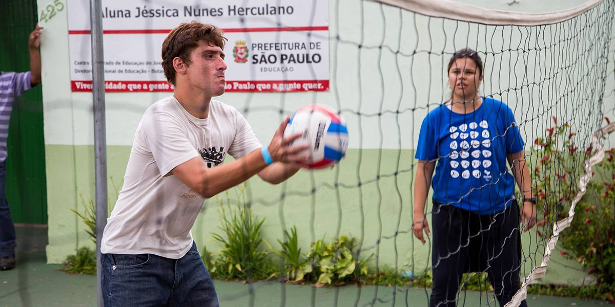Jovem segura bola de vôlei com as duas mãos diante da rede. Atrás, professora acompanha a jogada.