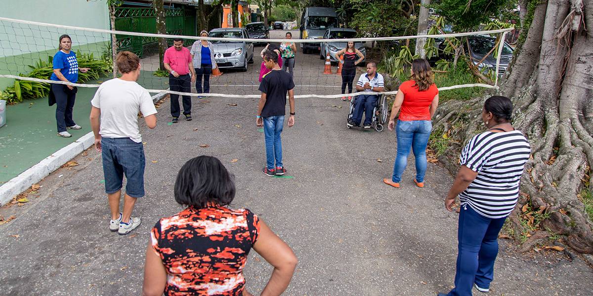 Dois times de adolescentes e adultos jogam vôlei de idosos em rua asfaltada.