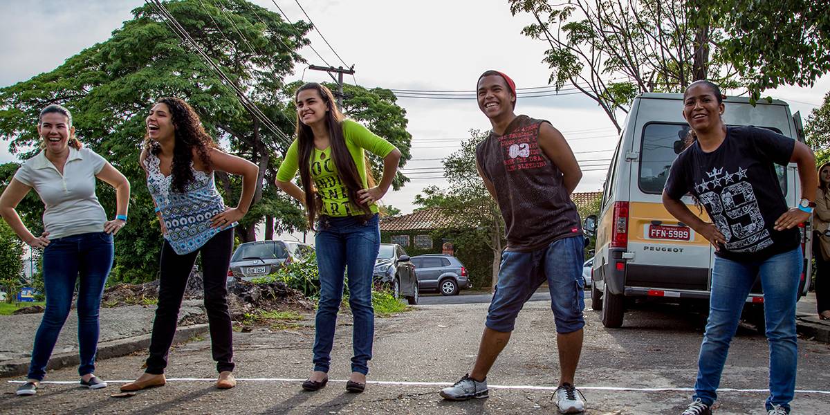 Um ao lado do outro, cinco adolescentes e adultos fazem alongamento girando seus troncos e com as mãos apoiadas na cintura.