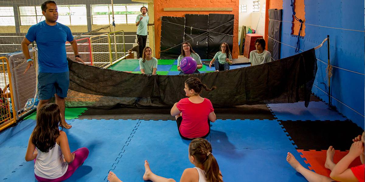 De pé, educador dá instruções para time de jovens em chão acolchoado durante partida de vôlei sentado.
