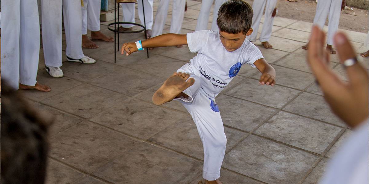Duas crianças jogam capoeira no centro de uma roda. Menina está agachada, com pés e mãos apoiados no chão e de pé, o garoto levanta uma das pernas.