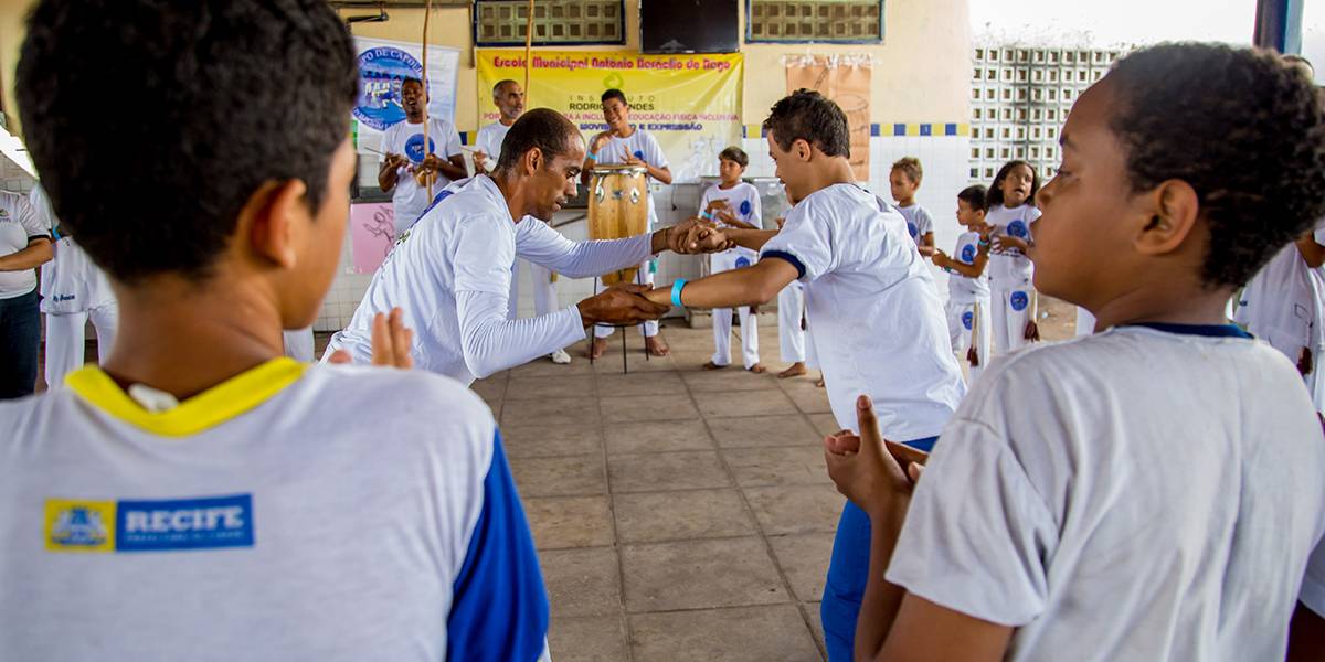 Professor ginga de mãos dadas com aluno com Síndrome de Down no centro de roda de capoeira.