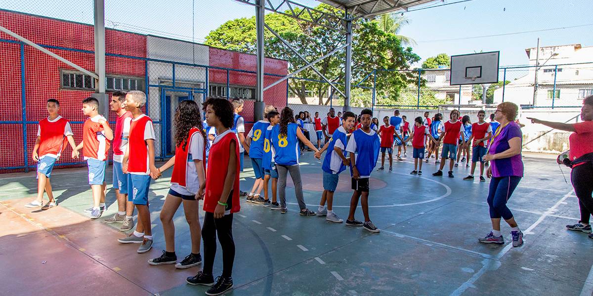 Estudantes de mãos dadas se posicionam em quadra como peças de uma mesa de pebolim. Eles estão divididos em dois times, caracterizados pelos coletes azuis e vermelhos. Há duas fileiras de cada time intercaladas. 