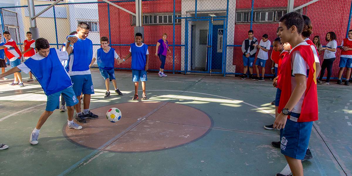Garoto de colete azul chuta bola de futebol para frente. Ele está de mãos dadas com colegas de time.