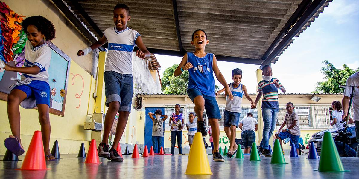Crianças sorridentes correm entre pequenos cones coloridos dispostos em fila no pátio escolar.