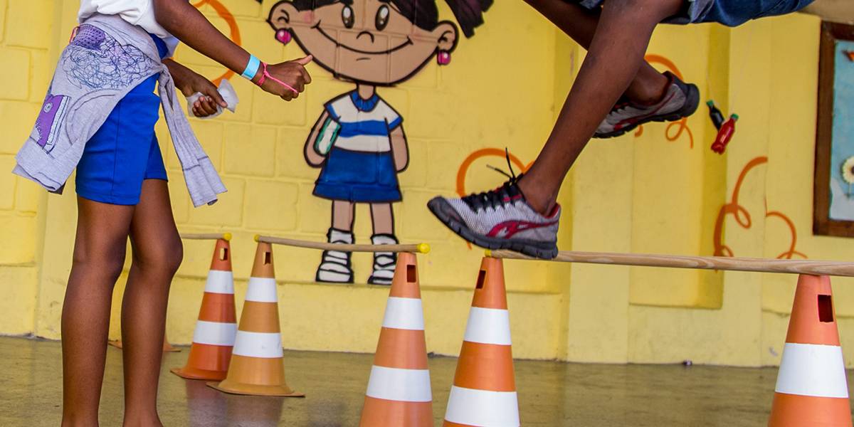 Garoto salta sobre obstáculo formado por um cabo de vassoura apoiado entre dois cones. Ao seu lado, colega acompanha o movimento e sorri.