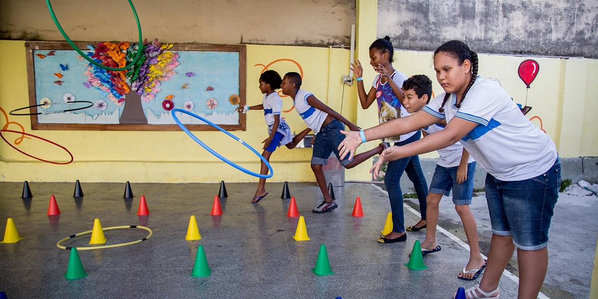Cinco crianças arremessam bambolês em direção a pequenos cones coloridos espalhados no chão do pátio.