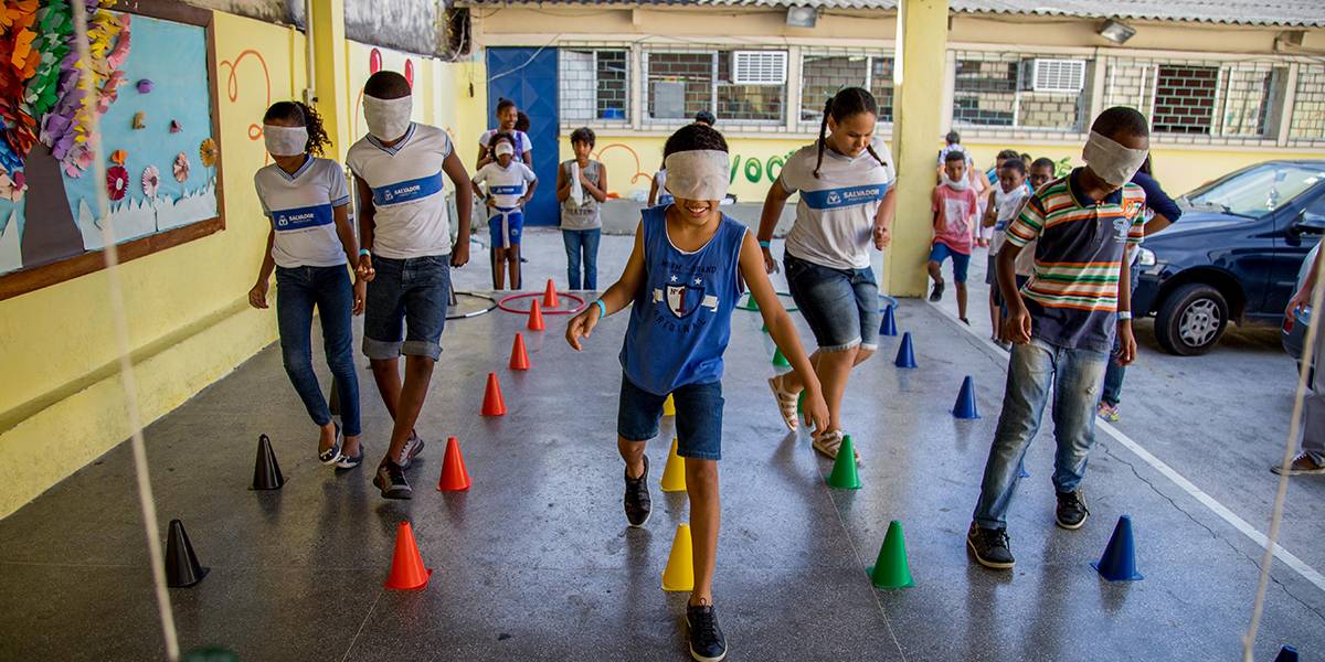 Meninas e meninos vendados caminham entre fileiras de cones coloridos no chão de um pátio. Raquel realiza a atividade sem a venda.