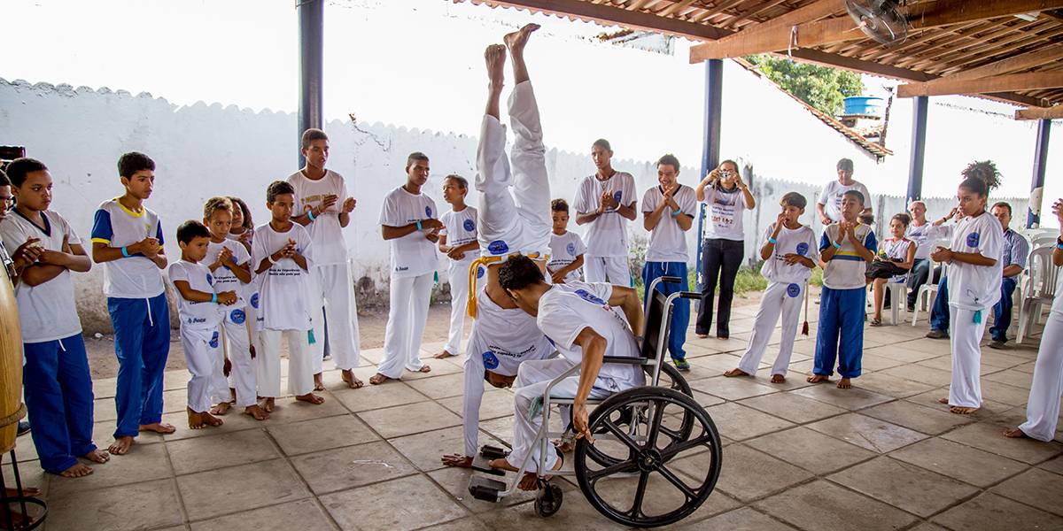Dois garotos jogam capoeira no centro de uma roda onde os demais batem palmas.  No centro, um deles dá uma pirueta apoiado com as mãos no chão e o outro, em uma cadeira de rodas, curva seu tronco para frente.