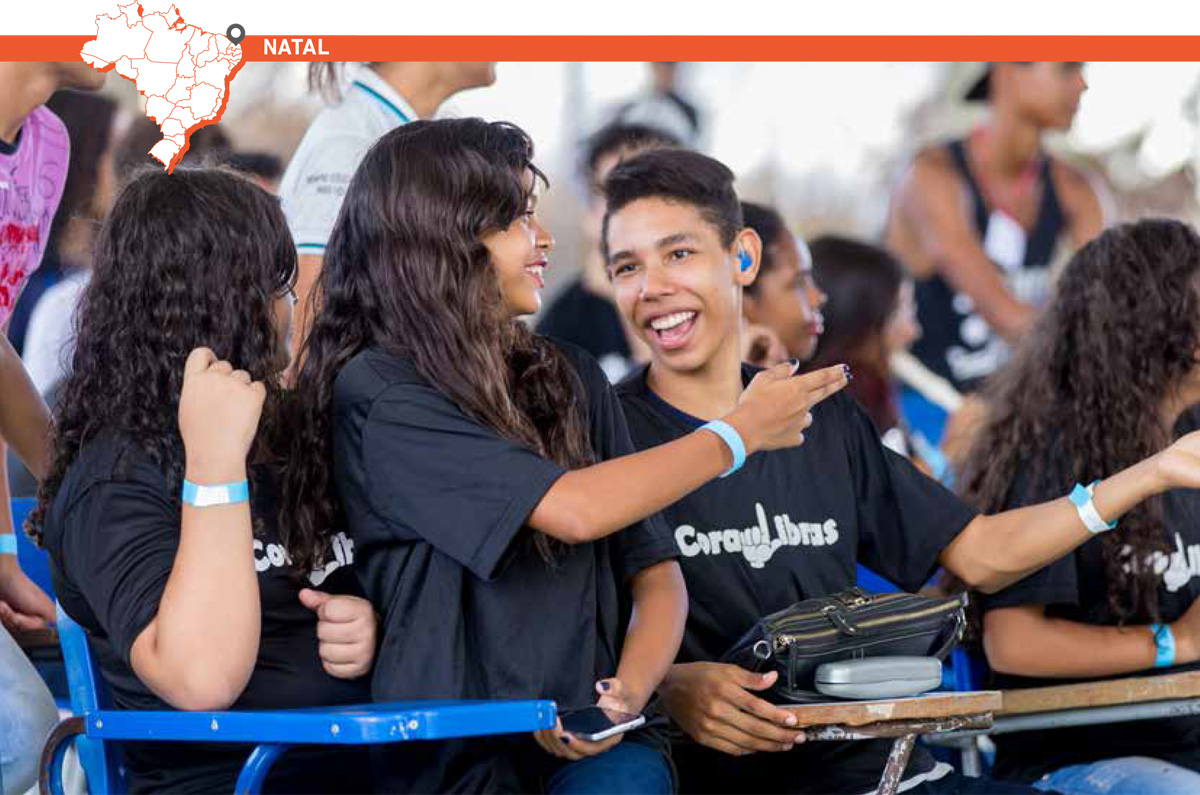 Adolescente com aparelho auditivo sorri e gesticula com uma colega. Eles vestem camisetas com estampa que diz “CoraLibras”. Acima da imagem, mapa do Brasil com marcação que destaca cidade de Natal.