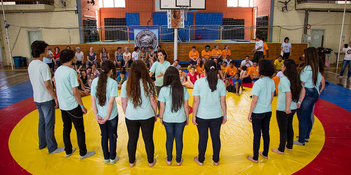 Em pé e descalços, grupo de adolescentes forma semicírculo entorno de educadora em chão acolchoado de quadra escolar. A frente deles, pessoas sentadas na arquibancada.
