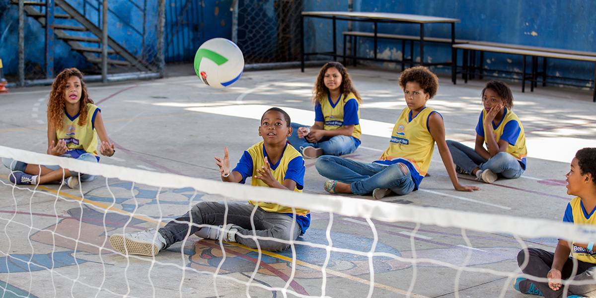 Menino sentado mantém as mãos abertas pronto para segurar a bola, ao fundo três meninas o observam