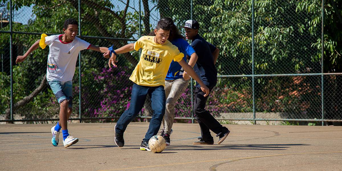 Alunos jogam futebol em dupla. Eles estão unidos pelo punho através de uma faixa de tecido.