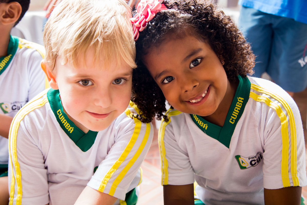 Menino loiro e menina negra sorriem, lado a lado, com as cabeças encostadas na altura da orelha.