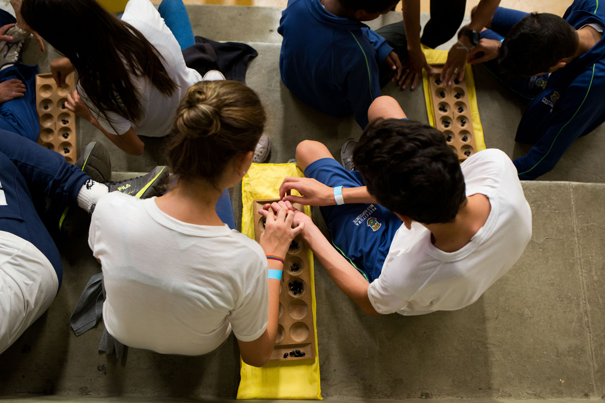 Menina e um menino com um tabuleiro de mancala vistos de cima. O objeto é feito de tábua com 12 orifícios onde alguns grãos de feijão estão depositados. 