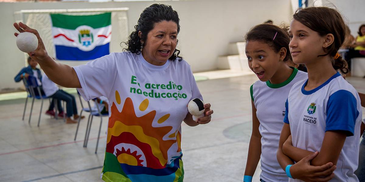 Professora segura bolinha enquanto orienta duas meninas.