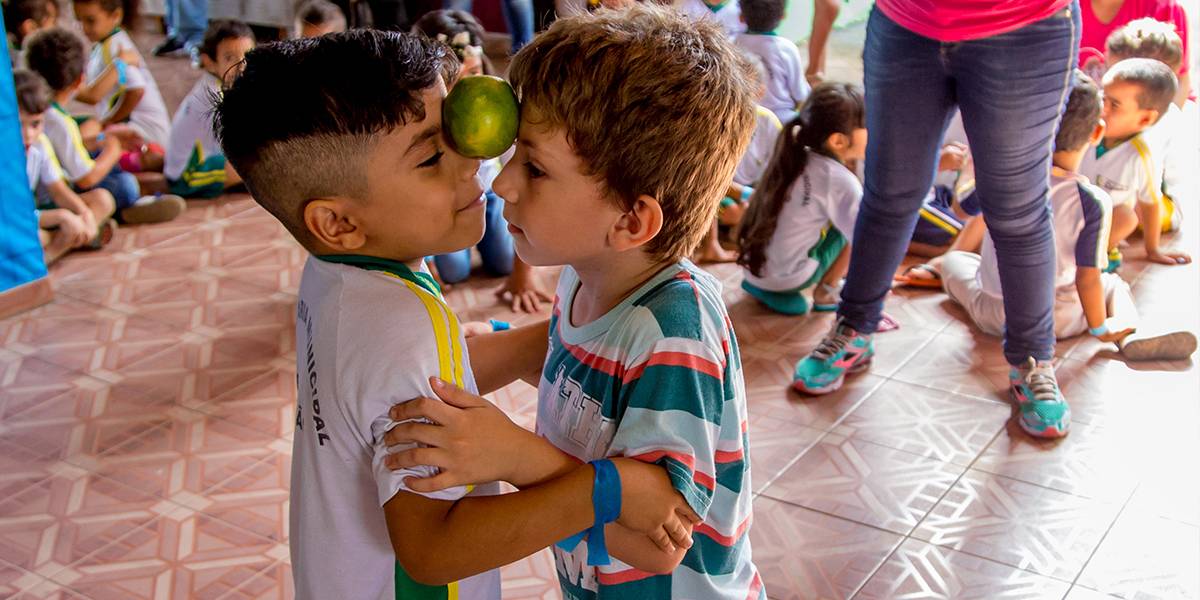 Dois meninos dançam abraçados com uma laranja equilibrada entre suas testas.
