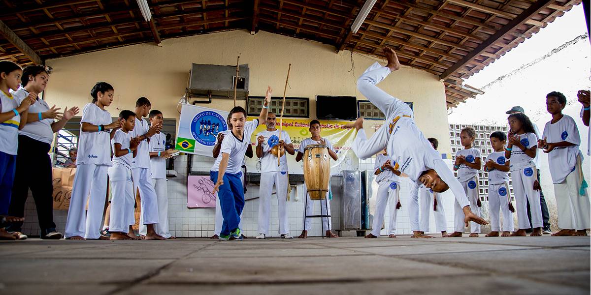 Professor faz pirueta ao lado de um aluno com deficiência intelectual. Eles estão no centro de uma roda de capoeira que reúne pessoas de diferentes idades que tocam instrumentos e batem palma. 